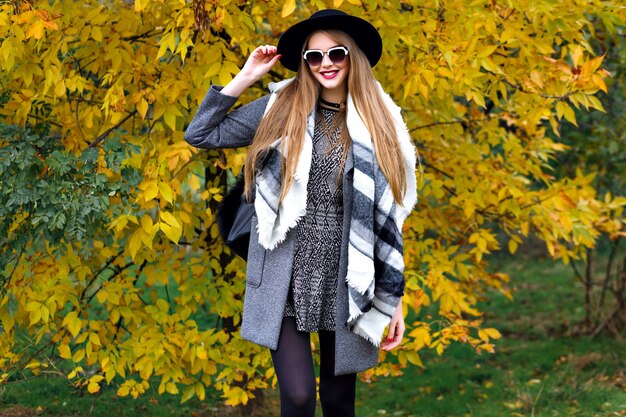 Retrato de moda de otoño de impresionante modelo elegante posando en el parque, hojas doradas y clima fresco, ropa de estilo callejero de lujo, maquillaje brillante, bufanda grande, mini vestido cubre abrigo y sombrero vintage.