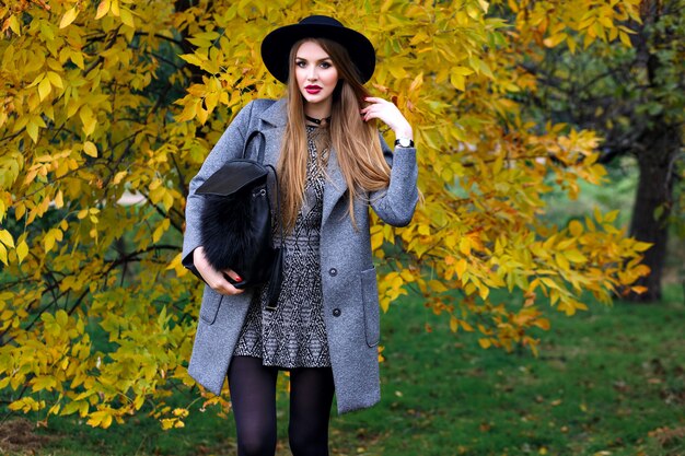 Retrato de moda otoño de mujer elegante glamour posando en el increíble parque de la ciudad, elegante abrigo, mochila y sombrero vintage.