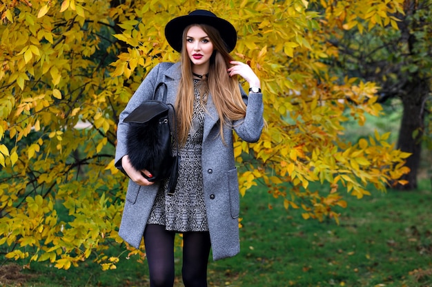Retrato de moda otoño de mujer elegante glamour posando en el increíble parque de la ciudad, elegante abrigo, mochila y sombrero vintage.