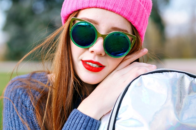 Retrato de moda positiva al aire libre de impresionante hermosa mujer jengibre, posando en el parque de la ciudad, con sombrero y clima hipster, gafas de sol de lujo de moda, juventud, vacaciones, labios rojos, colores pastel.