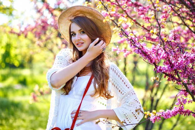 Retrato de moda soleado de primavera de mujer bonita rubia posando en el jardín floreciente, vestida con traje blanco boho y sombrero de paja.