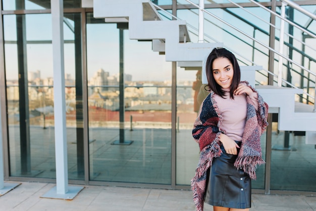 Retrato de moda sonriente joven morena en terraza en un día soleado en vistas a la gran ciudad. Sonriendo a un lado, éxito, tiempo de relajación, relajándose con la luz del sol.