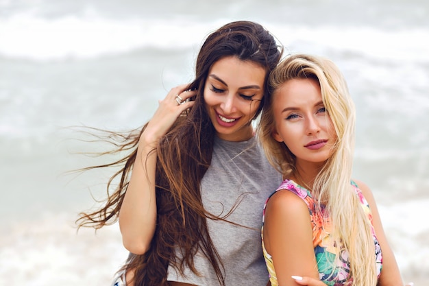 Retrato de moda de verano al aire libre de dos chicas bastante mejores amigos, posando cerca del océano, tiempo nublado con viento.