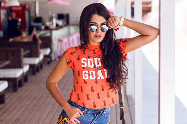 Retrato de moda de verano al aire libre de joven morena de pelo largo posando en el bar de la playa