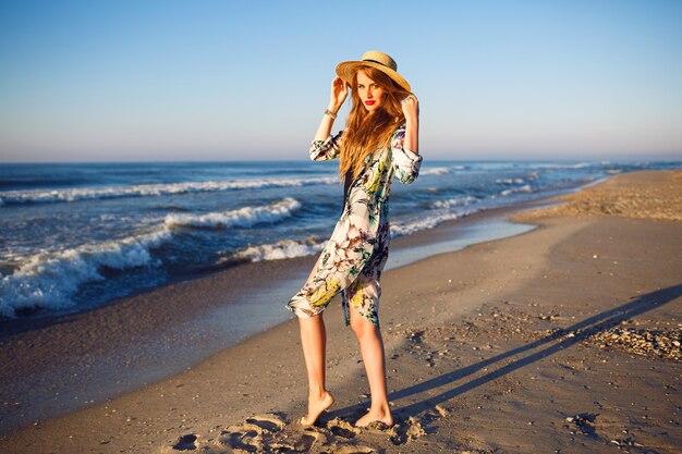 Retrato de moda de verano al aire libre de modelo rubia posando cerca del océano en la playa solitaria, colores tonificados, relajantes vacaciones de lujo