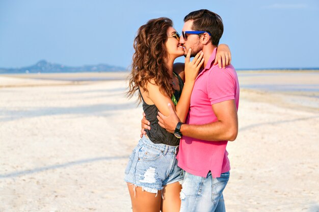 Retrato de moda de verano de joven pareja de hipsters muy elegantes enamorados abrazos y posando en la increíble playa de la isla, divirtiéndose solo, vistiendo ropa casual brillante y gafas de sol.