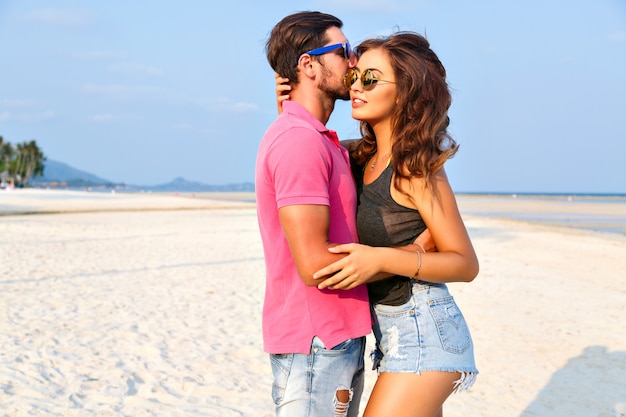 Retrato de moda de verano de joven pareja de hipsters muy elegantes enamorados abrazos y posando en la increíble playa de la isla, divirtiéndose solo, vistiendo ropa casual brillante y gafas de sol.