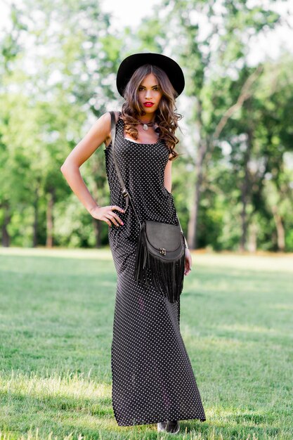 Retrato de moda de verano de mujer elegante con cabello ondulado perfecto en elegante elegante sombrero negro y maquillaje brillante posando en el parque.