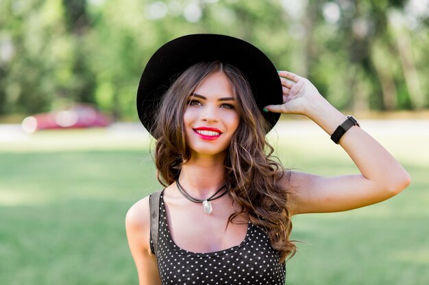 Retrato de moda de verano de mujer elegante con cabello ondulado perfecto en elegante elegante sombrero negro y maquillaje brillante posando en el parque.