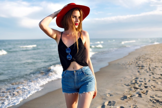 Retrato de moda de verano de mujer joven caminando sola cerca del océano, vacaciones en la playa, viaje solo, con gafas de sol de sombrero vintage y pantalones cortos de mezclilla, cuerpo delgado, amanecer, estilo de vida saludable.