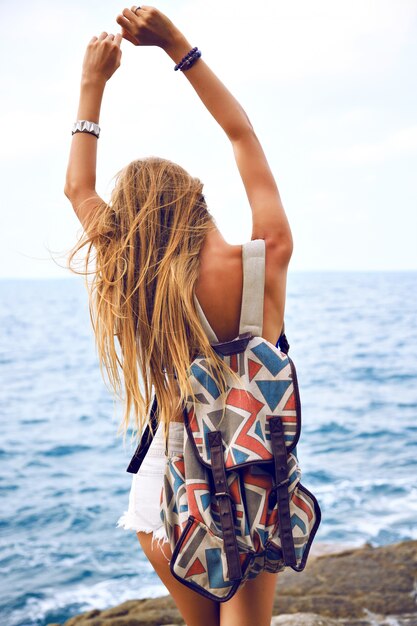 Retrato de moda de verano vaya mujer joven que viaja con mochila en verano, posando cerca del océano en un día lluvioso, viento triste.