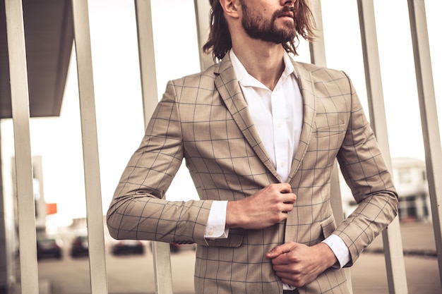 El retrato del modelo atractivo atractivo del hombre de negocios de la moda se vistió en el traje a cuadros elegante que presentaba cerca de la pared de ladrillo en el fondo de la calle.