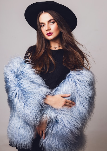 Retrato de modelo bastante joven en elegante abrigo mullido de invierno y sombrero negro posando