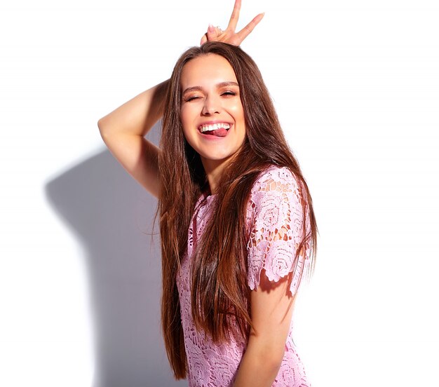 Retrato del modelo caucásico sonriente de la mujer morena hermosa en el vestido elegante del verano rosado brillante aislado en el fondo blanco. Mostrando su lengua y signo de paz