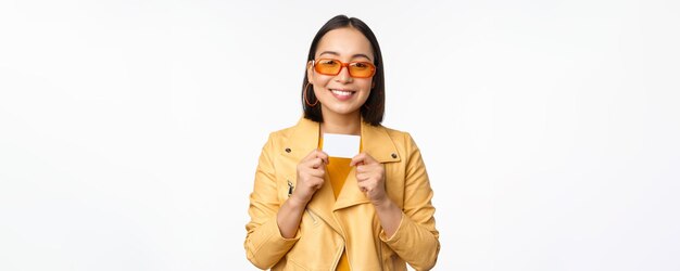 Retrato de una modelo femenina coreana sonriente con gafas de sol que muestra una tarjeta de crédito de pie sobre un fondo blanco