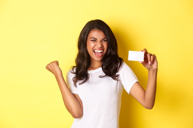 Retrato de modelo femenino afroamericano alegre mostrando tarjeta de crédito y gritando de alegría feliz