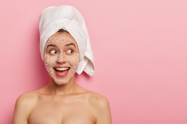 Retrato de modelo femenino feliz aplica exfoliante de sal marina en la cara, tiene expresión positiva, mira a un lado, tiene el cuerpo desnudo, usa una toalla después del baño