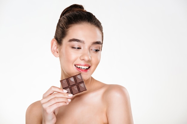 Retrato de modelo femenino lindo fresco con cabello oscuro comiendo barra de chocolate sin contar calorías
