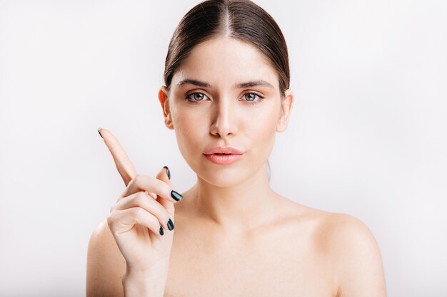 Retrato de modelo femenino sin maquillaje apuntando con el dedo índice hacia arriba en la pared aislada.