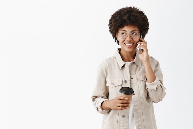 Retrato de modelo femenino de piel oscura amigable y atractivo hablador en gafas y camisa sosteniendo una taza de café de papel y hablando por teléfono celular mirando a la izquierda