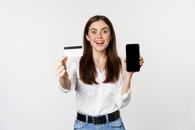 Retrato de modelo femenino que muestra la tarjeta de crédito con la pantalla del teléfono inteligente recomendando la aplicación standin...