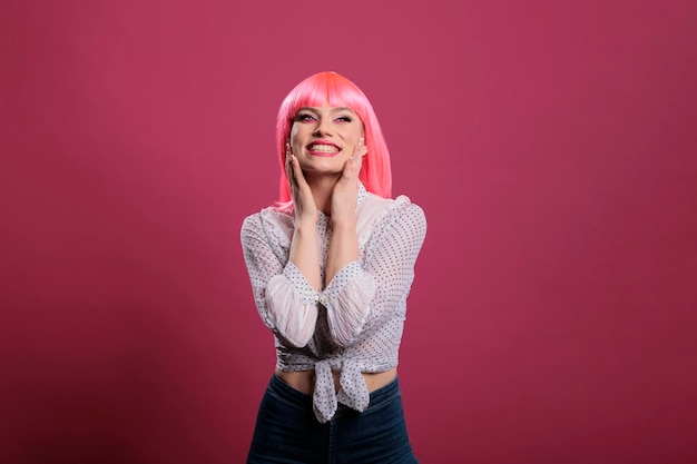 Retrato de modelo de glamour sonriendo y posando en cámara, sintiéndose feliz y positivo en el estudio. Mujer alegre despreocupada con peluca de pelo rosa y maquillaje elegante haciendo movimientos sensuales y atractivos.