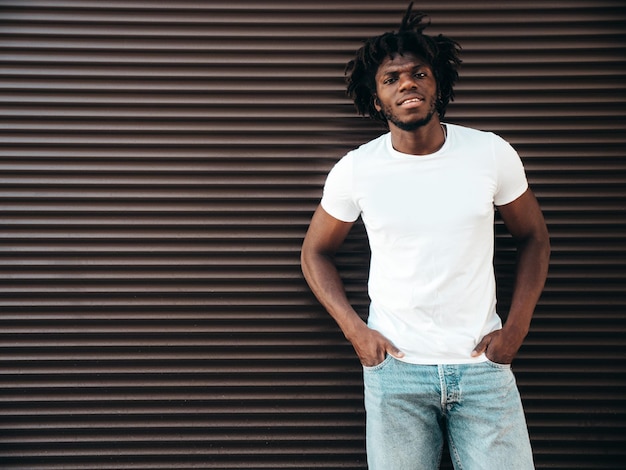 Retrato de modelo hipster guapo Un hombre africano sin afeitar vestido con camiseta blanca de verano y jeans Hombre de moda con peinado de rastas posando cerca de la pared de la persiana enrollable en la calle