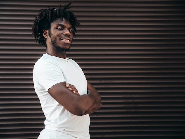 Retrato de modelo hipster guapo sonriente Hombre africano sin afeitar vestido con camiseta blanca de verano y jeans Hombre de moda con peinado de rastas posando en el fondo de la calle
