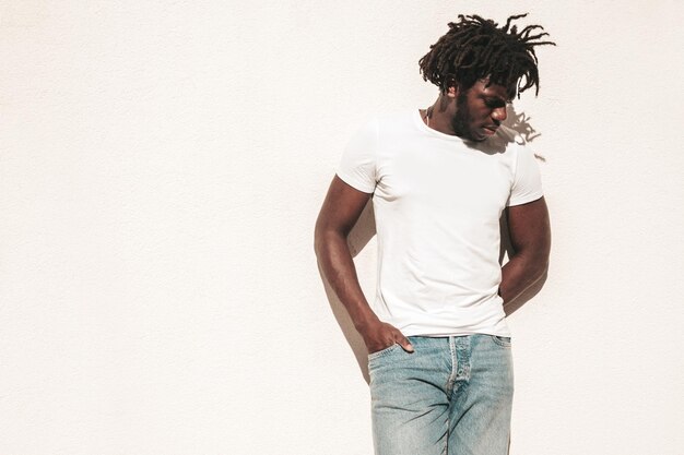 Retrato de modelo hipster guapo sonriente Hombre africano sin afeitar vestido con camiseta blanca de verano y jeans Hombre de moda con peinado de rastas posando en el fondo de la calle