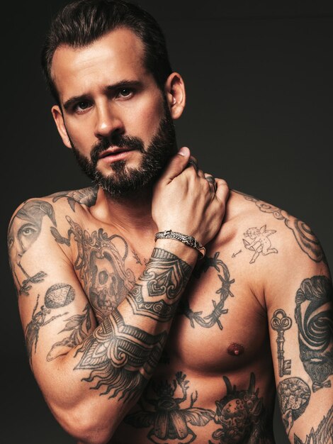 Retrato de modelo lambersexual guapo confiado con estilo hipster Hombre moderno sexy Torso desnudo con tatuajes Moda hombre posando en estudio sobre fondo oscuro