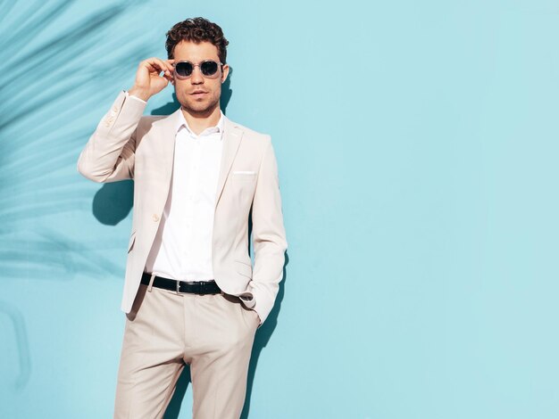 Retrato de modelo lambersexual guapo con estilo hipster Hombre moderno sexy vestido con traje elegante blanco Hombre de moda posando en el estudio cerca de la pared azul en gafas de sol