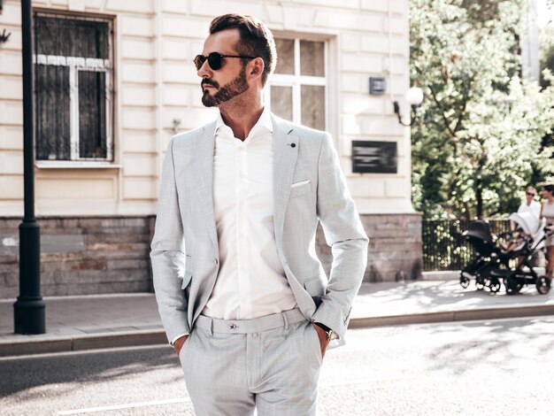 Retrato de un modelo lambersexual guapo y seguro de sí mismo con estilo hipster Hombre moderno vestido con un elegante traje blanco Hombre de moda posando en el fondo de la calle en la ciudad de Europa al atardecer Con gafas de sol