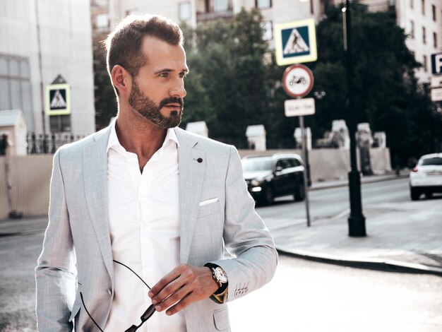 Retrato de un modelo lambersexual guapo y seguro de sí mismo con estilo hipster Hombre moderno vestido con un elegante traje blanco Hombre de moda posando en el fondo de la calle en la ciudad de Europa al atardecer Con gafas de sol