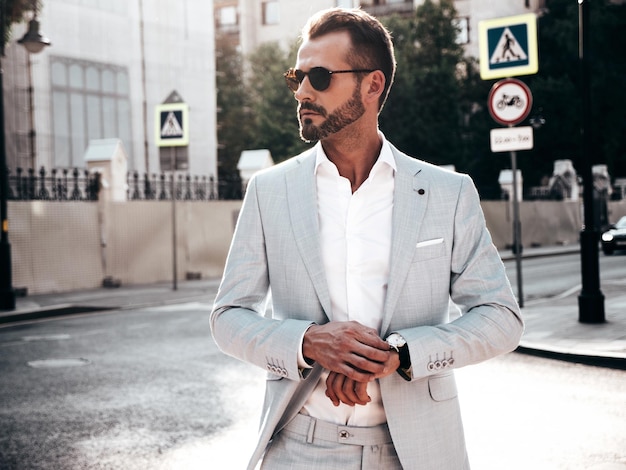 Retrato de un modelo lambersexual guapo y seguro de sí mismo con estilo hipster Hombre moderno vestido con un elegante traje blanco Hombre de moda posando en el fondo de la calle en la ciudad de Europa al atardecer Con gafas de sol