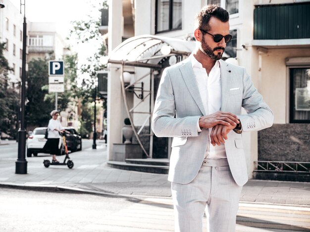 Retrato de un modelo lambersexual guapo y seguro de sí mismo con estilo hipster Hombre moderno vestido con un elegante traje blanco Hombre de moda posando en el fondo de la calle en la ciudad de Europa al atardecer Con gafas de sol