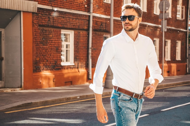 Retrato de un modelo lambersexual de hipster elegante y seguro de sí mismo Hombre moderno vestido con camisa blanca Hombre de moda posando en el fondo de la calle con gafas de sol Al aire libre al atardecer