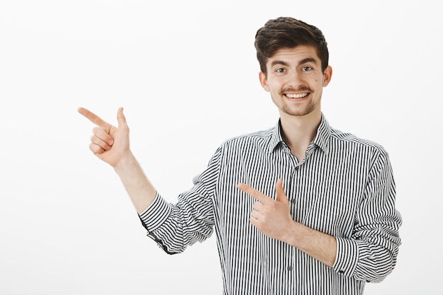 Retrato de modelo masculino europeo divertido coqueto con bigote y barba en camisa a rayas, apuntando hacia la izquierda con gestos de pistola y sonriendo ampliamente, invitando a una linda mujer a seguir hablando en el bar