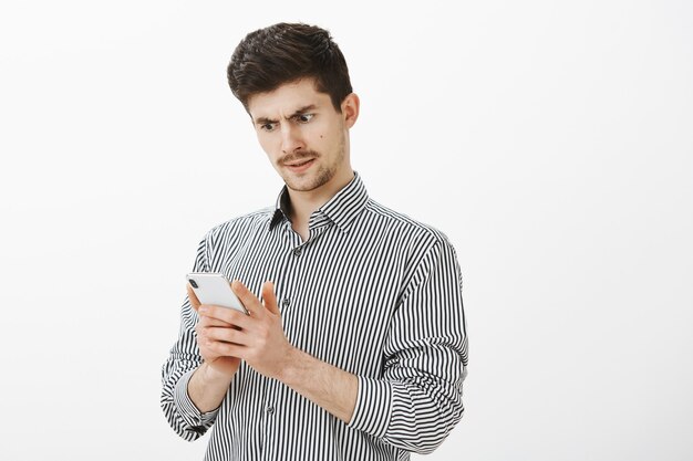 Retrato de un modelo masculino europeo ordinario confundido intenso con bigote, mirando interrogado en la pantalla del teléfono inteligente, recibiendo un mensaje extraño o sin tener ni idea de dónde se fue el dinero de la cuenta bancaria