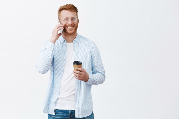 Retrato del modelo masculino europeo tranquilo y relajado despreocupado con cerdas y pelo rojo, sosteniendo una taza de café de papel, sosteniendo el teléfono inteligente cerca de la oreja