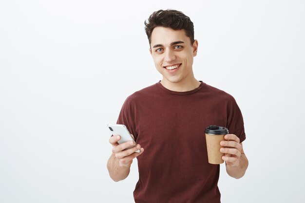 Retrato de modelo masculino guapo sin preocupaciones en camiseta roja con smartphone y taza de café