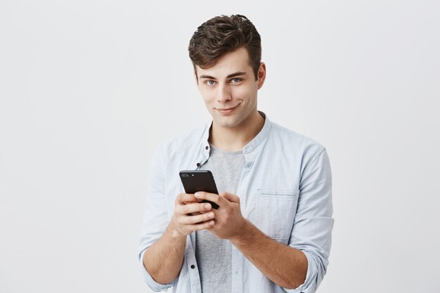 Retrato del modelo masculino hermoso atractivo que lleva la camisa azul que sostiene el teléfono elegante moderno usando la conexión a internet de alta velocidad, enviando mensajes de texto a sus amigos. Tecnología moderna y comunicación.
