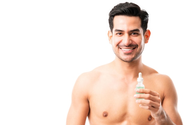 Retrato de un modelo masculino joven feliz que sostiene suero hidratante de ácido hialurónico mientras hace contacto visual con fondo blanco