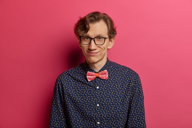 Retrato de modelo masculino positivo divertido con expresión complacida, viste una camisa elegante, gafas transparentes, está de buen humor, sale a la cita, espera a la novia, posa contra la pared rosa