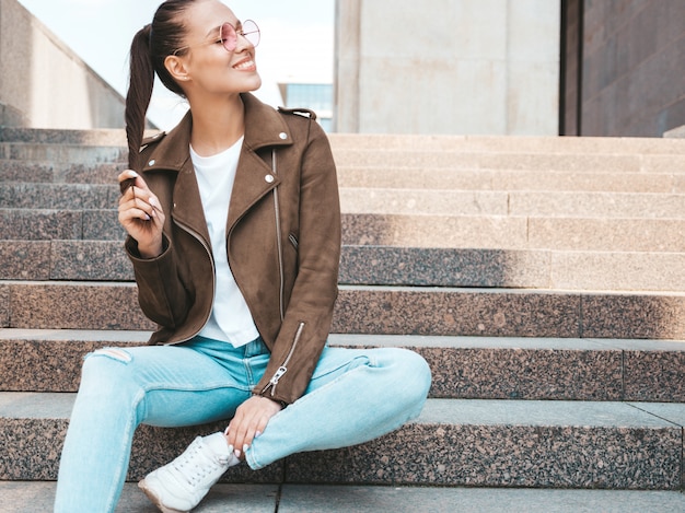 El retrato de la modelo morena sonriente hermosa se vistió en ropa de la chaqueta y de los vaqueros del inconformista del verano. Moda niña sentada en pasos en el fondo de la calle. Mujer divertida y positiva en gafas de sol redondas