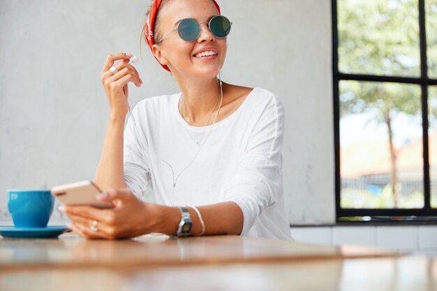 Retrato de modelo de mujer alegre se pone auriculares, disfruta de una canción perfecta o música favorita, conectado a un teléfono celular moderno, se sienta a la mesa con una taza de café contra el interior del café Concepto de personas y descanso