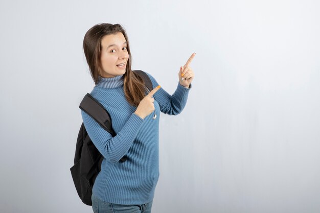 Retrato de un modelo de mujer joven con mochila apuntando hacia arriba con los dedos índices.