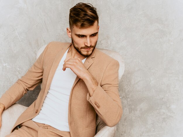 Retrato del modelo sonriente hermoso del hombre de negocios del inconformista que lleva el traje beige casual. sentado en una silla en el interior