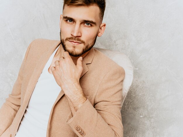 Retrato del modelo sonriente hermoso del hombre de negocios del inconformista que lleva el traje beige casual. sentado en una silla en el interior