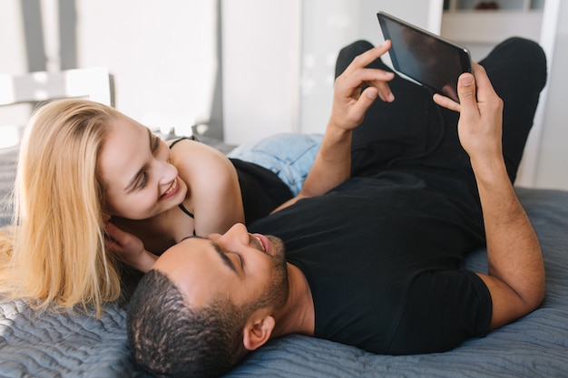 Retrato momentos encantadores de la feliz pareja de linda mujer joven y chico guapo navegando en internet en tableta en cama en apartamento moderno. Divirtiéndose, familia joven, amor, alegría, sonriendo