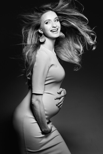 Retrato monocromático de hermosa mujer embarazada feliz vestida abrazando el vientre y sonriendo Positividad y mujer elegante posando mirando a la cámara Concepto de moda de embarazo Efecto ventoso